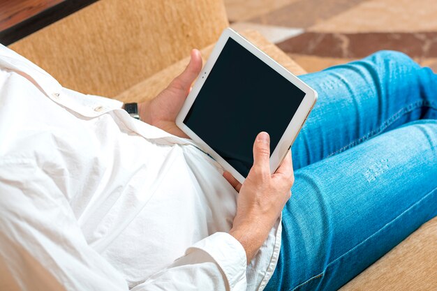 Joven en camisa blanca y jeans azul trabajando y mirando en tableta con pantalla negra en la oficina en casa en un acogedor sofá beige