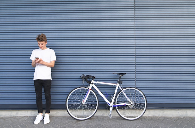 Joven con una camisa blanca de pie cerca de la bicicleta de carretera y utiliza el teléfono inteligente