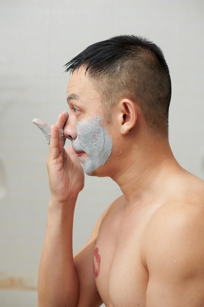 Joven sin camisa mirando el espejo del baño al aplicar una máscara de arcilla limpiadora de poros profundos