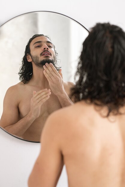 Joven sin camisa mirando su barba en el espejo mientras se va a afeitar después de bañarse por la mañana