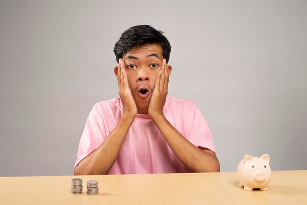 El joven con camisa rosa se sorprende mientras mira la cámara frente a las monedas y la alcancía rosa