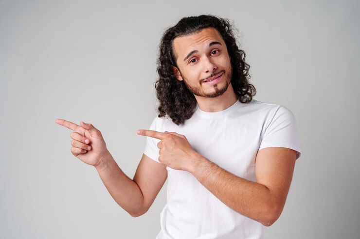 Foto un joven con una camiseta blanca y vaqueros señalando con confianza mientras sonríe en un entorno de estudio