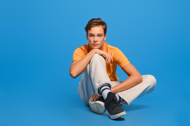 Joven con camiseta naranja y pantalones de chándal blancos sentado en el suelo del estudio y levantando la barbilla sobre fondo azul