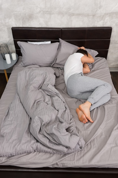 Joven cansado en pijama durmiendo sin manta en la elegante cama en colores grises y junto a la mesita de noche con velas en un dormitorio de estilo loft