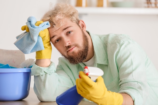 Joven cansado con productos de limpieza en la cocina