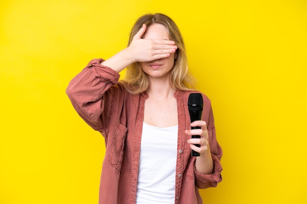 Joven cantante caucásica recogiendo un micrófono aislado de fondo amarillo cubriendo los ojos con las manos No quiero ver algo