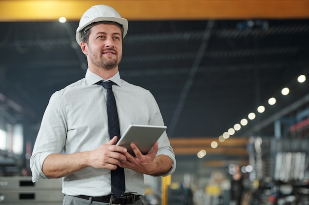 Joven capataz elegante exitoso con touchpad trabajando con datos y controlando el trabajo de la máquina industrial