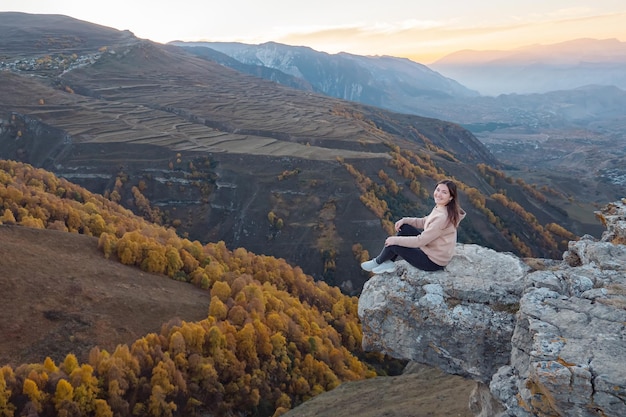 Foto una joven con capucha marrón se sienta en el borde superior de una colina contra un paisaje montañoso con naranjos y campos en otoño