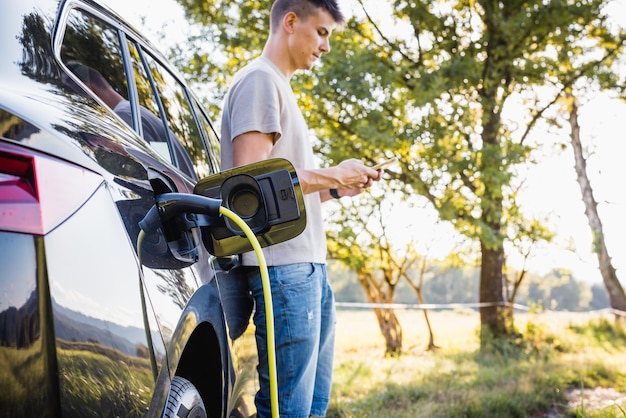 Joven cargando un auto eléctrico estacionado en el área natural y ajustando una aplicación de carga de vehículos eléctricos