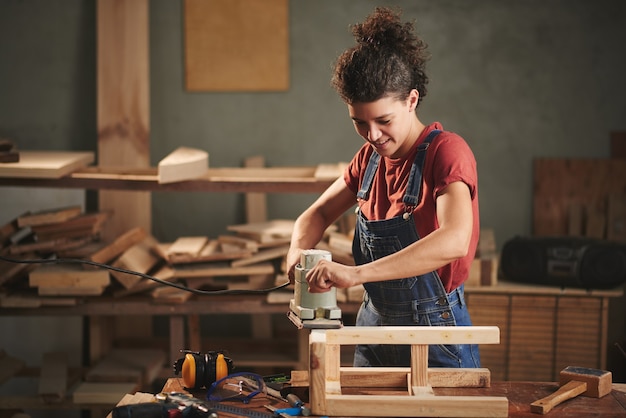 Joven carpintero bastante femenino alisando cuidadosamente el taburete de madera con lijadora de banda eléctrica y sonriendo