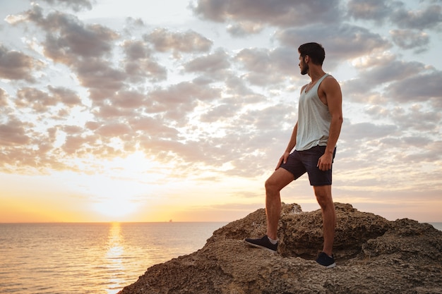 Joven casual de pie sobre la roca de la montaña junto al mar y mirando al atardecer