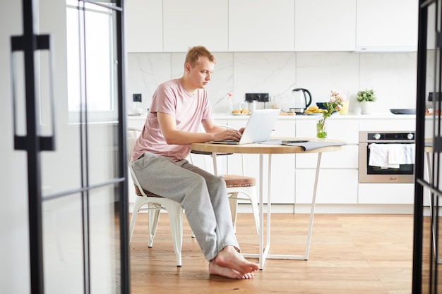 Joven casual sentado en la mesa en la cocina frente a la computadora portátil