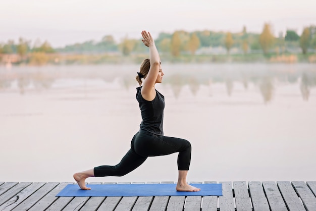 Una joven caucásica de 36 años practica yoga al aire libre cerca de un río en un muelle de madera en la mañana