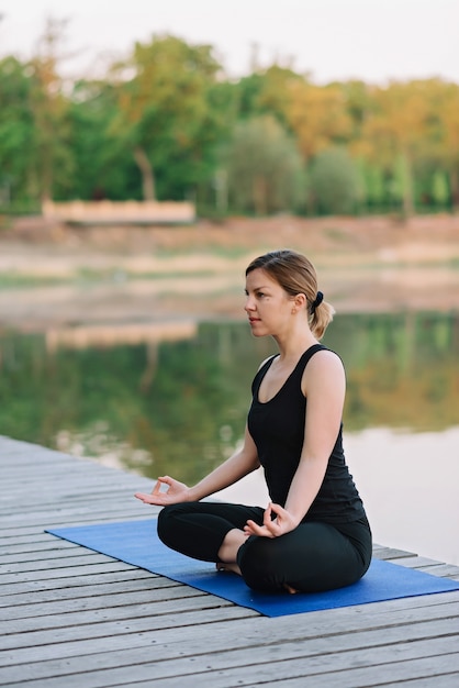 Una joven caucásica de 36 años practica yoga en la posición de loto al aire libre cerca del río en un muelle de madera en la mañana