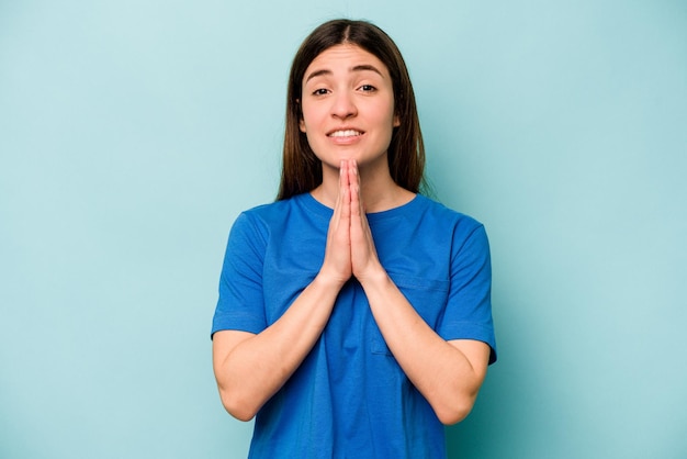 Una joven caucásica aislada de fondo azul tomándose de la mano para orar cerca de la boca se siente segura