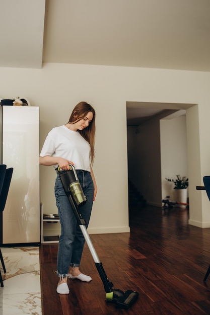 Una joven caucásica aspira el suelo de la cocina con una aspiradora vertical inalámbrica o una escoba eléctrica La criada está limpiando la sala de cocina del albergue Una mujer blanca limpia el interior