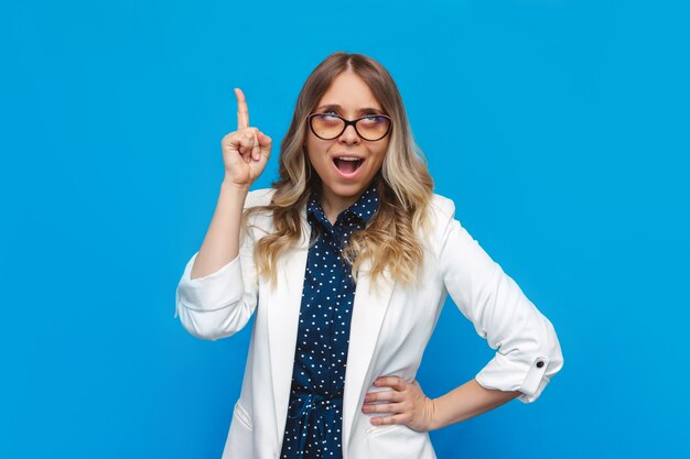 Una joven caucásica bastante inteligente mujer de negocios rubia con una chaqueta blanca y gafas mira hacia arriba gesticulando con la mano y el dedo hacia arriba se siente emocionada con una buena idea aislada en una pared de color azul brillante