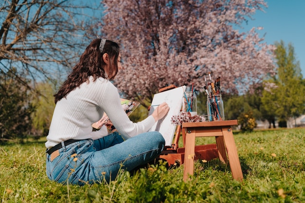 Joven caucásica con cabello ondulado pintando un lienzo con pinturas de color rosa pastel mientras está de pie en la naturaleza de un parque, frente a un almendro rosa gigante.