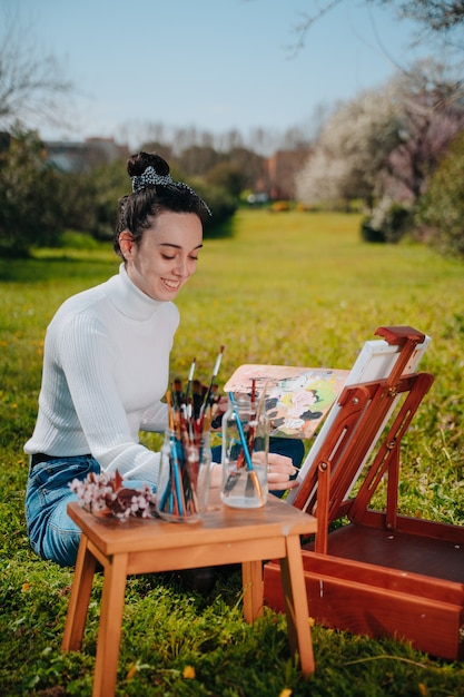 Joven caucásica con cabello ondulado pintando un lienzo con pinturas de color rosa pastel mientras está de pie en la naturaleza de un parque