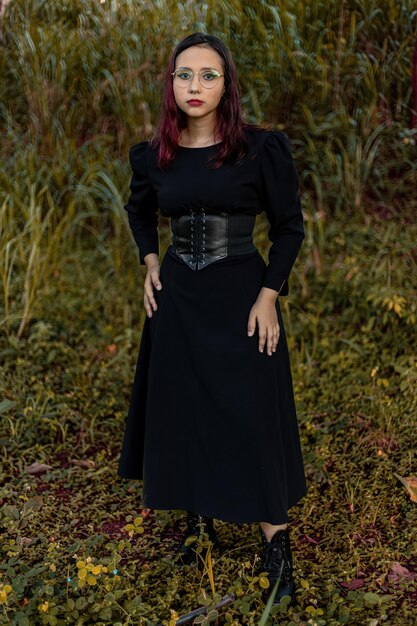Joven caucásica de cabello oscuro con rayas rojas vestidas de negro con cinturón de corsé y gafas posando para una sesión de belleza femenina