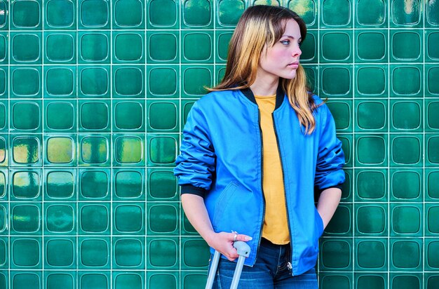 Joven caucásica en chaqueta azul esperando apoyado en una pared verde con una maleta