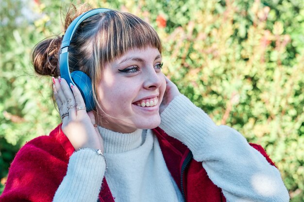 Joven caucásica en una chaqueta roja que está escuchando música con auriculares azules en el parque