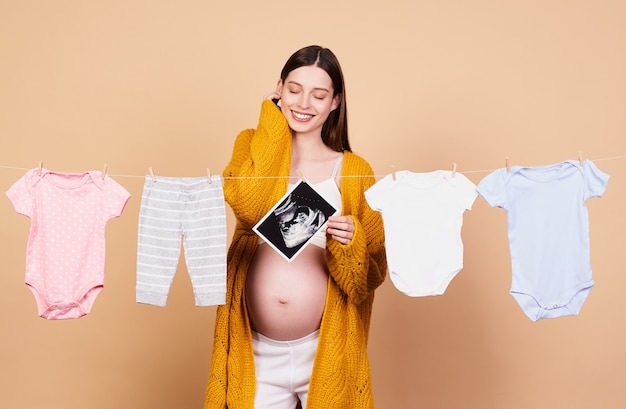 Una joven caucásica dulce, soñadora, gentil y romántica con cabello largo y oscuro, con un cárdigan de punto amarillo, se prepara para el próximo nacimiento, recolecta ropa de bebé, se regocija.