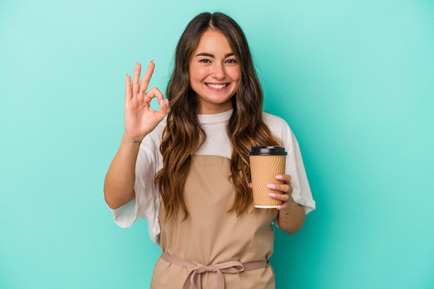 Joven caucásica empleada de una tienda sosteniendo un café para llevar aislado en un fondo azul alegre y confiada mostrando un gesto correcto.