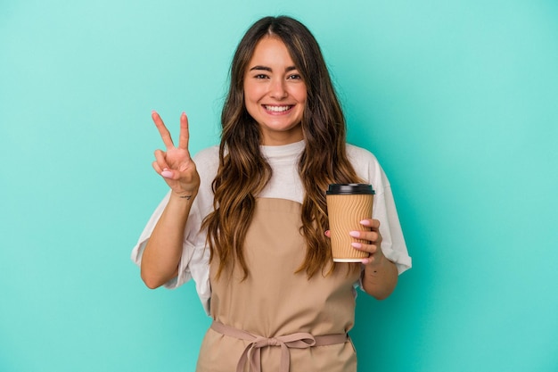 Joven caucásica empleada de una tienda sosteniendo un café para llevar aislado en un fondo azul que muestra el número dos con los dedos.