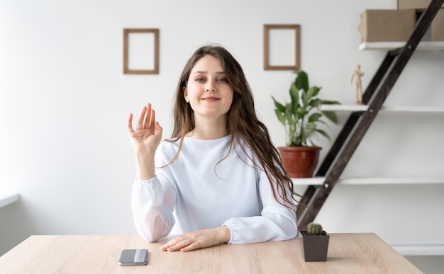 Una joven caucásica está sentada en su escritorio en casa, mira la cámara y saluda alegremente Comunicación en línea y concepto de chat de video vista de cámara web