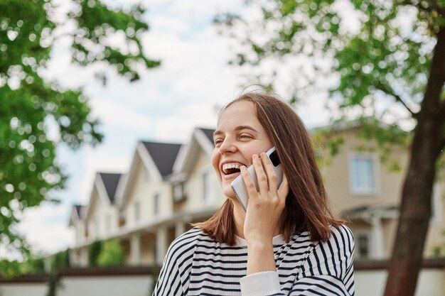 Una joven caucásica feliz hablando con un celular en la calle con edificios en el fondo sosteniendo un teléfono móvil riéndose y teniendo una agradable conversación con amigos