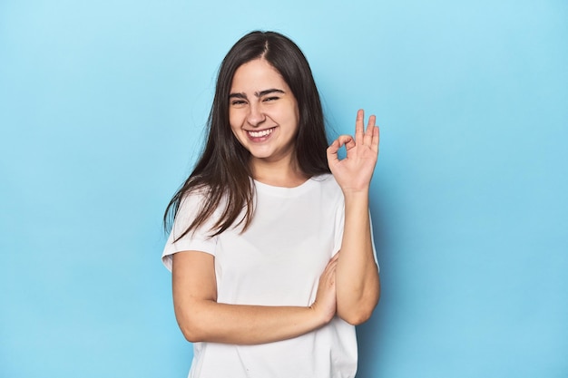 Una joven caucásica con fondo azul guiña un ojo y hace un buen gesto con la mano