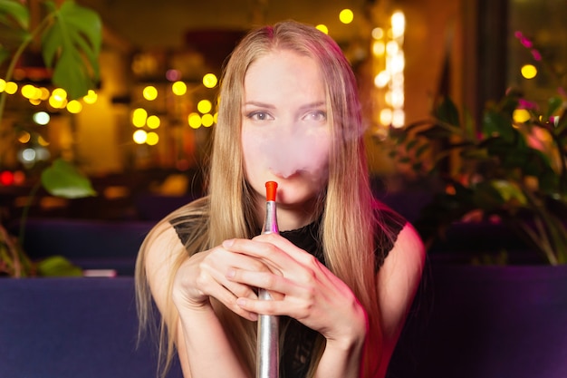Una joven caucásica fuma un narguile o una shisha en el club nocturno o en el bar.