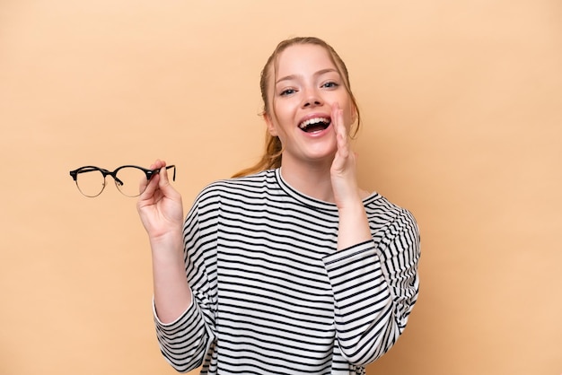 Joven caucásica con gafas aisladas de fondo beige gritando con la boca abierta