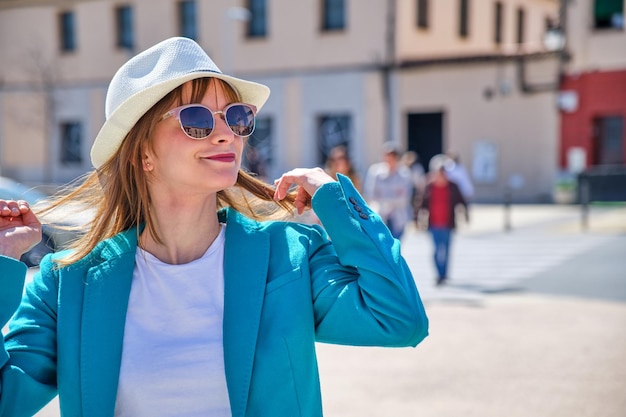 Joven caucásica con gafas de sol y sombrero blanco tocándose el pelo caminando en un día soleado