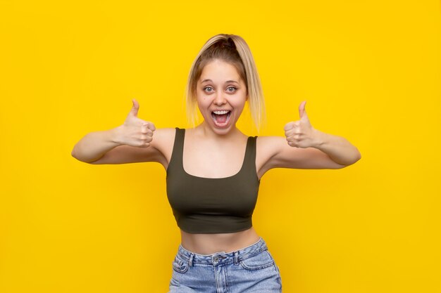 Una joven caucásica hermosa feliz emocionada encantada sonriente mujer rubia con una cola de caballo que muestra el pulgar hacia arriba como gesto con las dos manos aisladas en una pared de color amarillo brillante