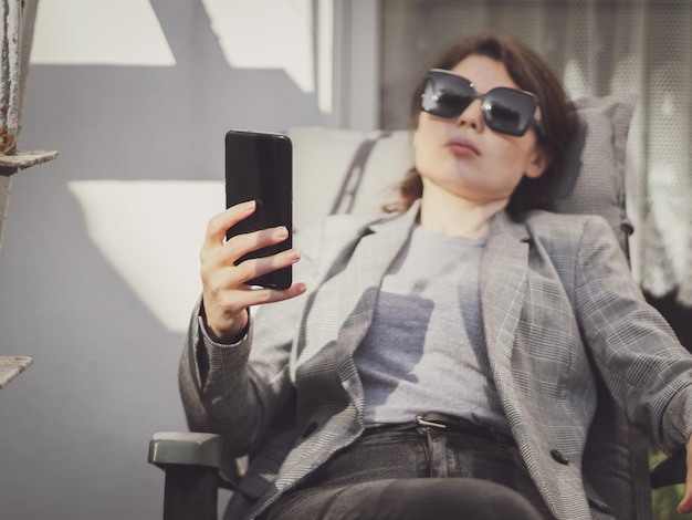 Una joven caucásica hermosa sostiene un teléfono inteligente frente a ella mientras se sienta en una silla de jardín