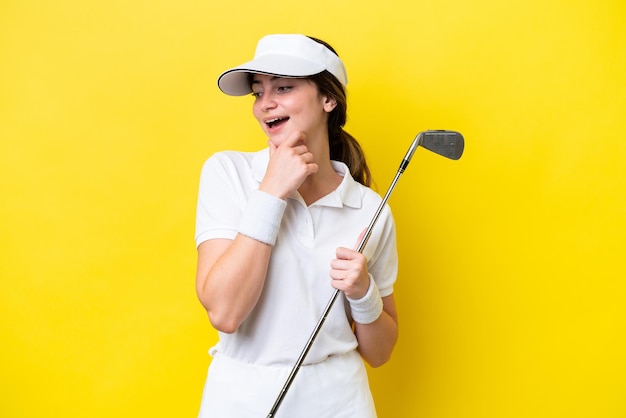 Una joven caucásica jugando al golf aislada de fondo amarillo mirando hacia un lado y sonriendo