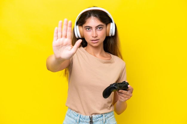 Una joven caucásica jugando con un controlador de videojuegos aislado en un fondo amarillo haciendo un gesto de parada