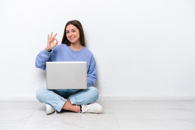 Una joven caucásica con una laptop sentada en el suelo aislada de fondo blanco mostrando el signo de ok con los dedos