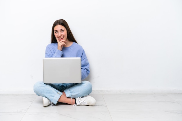 Una joven caucásica con una laptop sentada en el suelo aislada de fondo blanco pensando en una idea mientras mira hacia arriba