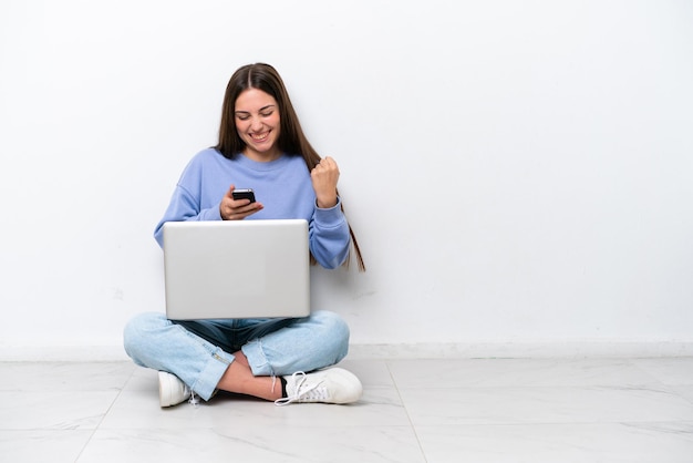 Una joven caucásica con una laptop sentada en el suelo aislada de fondo blanco sorprendida y enviando un mensaje
