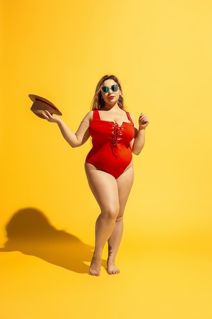 Joven caucásica modelo femenino de talla grande preparándose para resort de playa en la pared amarilla. Mujer en traje de baño rojo, sombrero y gafas de sol. Concepto de verano, fiesta, cuerpo positivo, igualdad y relajación.