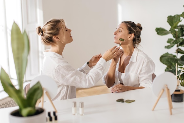 Una joven caucásica le muestra a su madre cómo usar el rodillo de jade mientras se sienta a la mesa en la habitación. Concepto de cosmética natural, alisado de arrugas