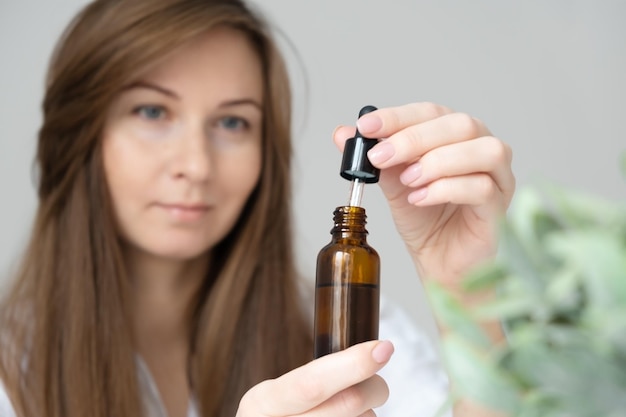 Joven caucásica mujer morena de pelo largo desenfocada haciendo procedimientos de belleza para el cuidado de la piel en el hogar, sosteniendo una botella de vidrio con suero, aceite, cosméticos bionaturales para el cabello o la cara en el hogar, primer plano