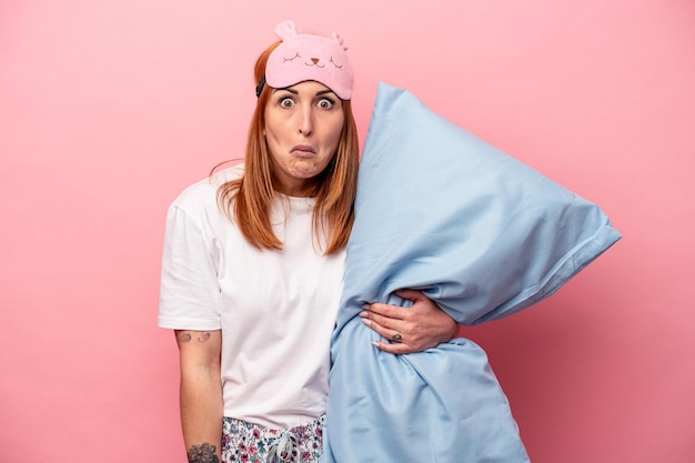 Una joven caucásica con un pijama sosteniendo una almohada aislada en un fondo rosado se encoge de hombros y abre los ojos confundidos.