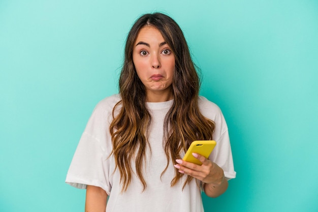 Una joven caucásica que sostiene un teléfono móvil aislado de fondo azul se encoge de hombros y abre los ojos confundida.