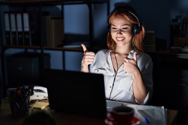 Una joven caucásica que trabaja en la oficina por la noche muestra el éxito haciendo un gesto positivo con la mano, pulgares arriba sonriendo y feliz. expresión alegre y gesto ganador.