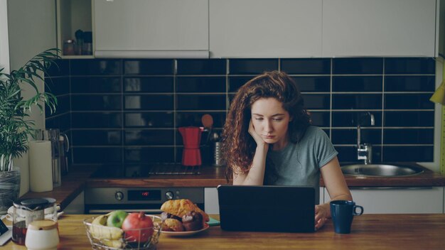 Una joven caucásica rizada y pensativa está sentada en la cocina adentro frente a una laptop tomando café y trabajando, echa un vistazo a la ventana