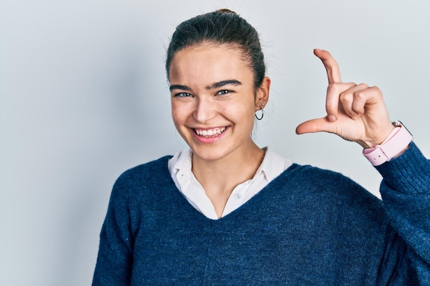 Joven caucásica con ropa informal sonriendo y confiada gesticulando con la mano haciendo un signo de tamaño pequeño con los dedos mirando y el concepto de medida de la cámara